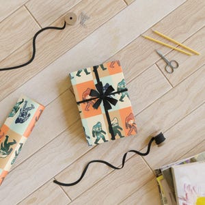 May include: A rectangular gift box wrapped in paper featuring a repeating grid pattern of Bigfoot illustrations in orange, teal, and cream colors, tied with a black ribbon. A roll of matching paper, black ribbon, two yellow pencils, and scissors are present.