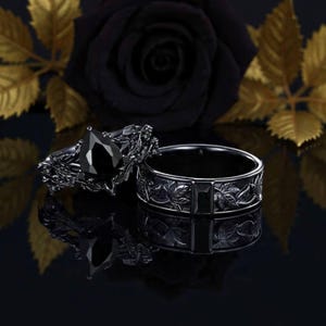 May include: Two black rings. One ring features a large, pear-shaped black gemstone. The other ring is a band with a leaf pattern and a rectangular black stone. The background includes a black rose and gold leaves.