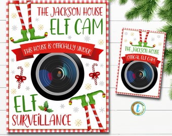 May include: A festive Christmas-themed sign with the text "THE JACKSON HOUSE ELF CAM" and "ELF SURVEILLANCE". The design includes a camera lens, elf legs, candy canes, and snowflakes. A smaller version of the sign is also visible.