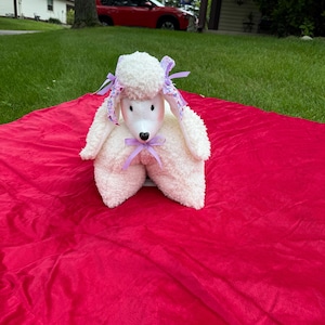 Peut inclure: Un jouet en peluche en forme de caniche blanc, avec un visage et des oreilles roses, orné de rubans violets. Le jouet est posé sur une couverture rouge vif, créant un contraste saisissant. En arrière-plan, une pelouse verte et une voiture rouge.