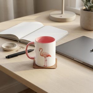 Könnte beinhalten: Ein weißer Keramikbecher mit einer Illustration eines rosa Flamingos auf der Vorderseite, einem einfarbig rosa Innenraum und einem passenden rosa Henkel. Der Becher steht auf einem braunen quadratischen Untersetzer und hat ein schlichtes Design.