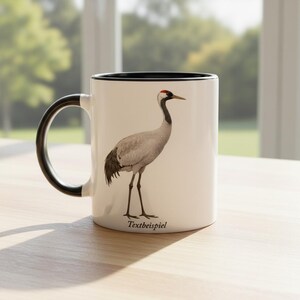 Peut inclure: Un mug en céramique blanche avec une anse et un bord intérieur noirs, orné d'une illustration détaillée d'une grue grise avec une couronne rouge sur la tête. Le mot Textbeispiel est imprimé en police serif noire au bas du mug.