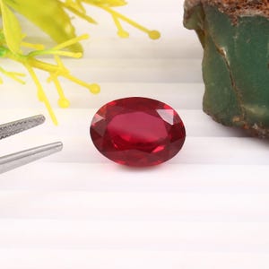 May include: A faceted oval-shaped gemstone with a deep red color. The stone features a polished surface with multiple geometric cuts that reflect light. A pair of silver-toned metal tweezers is positioned to the left of the gemstone.