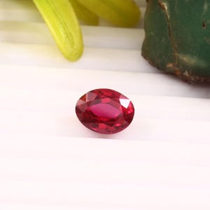 May include: A faceted, oval-shaped, deep red gemstone. The gem is set against a white background, with hints of green and yellow foliage in the periphery. The stone's facets reflect light, showcasing its brilliance.