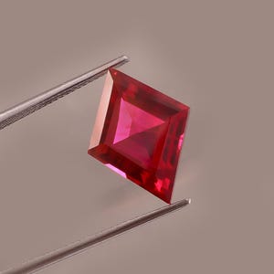 May include: A kite-shaped, faceted gemstone with a vibrant, deep red colour. The stone is held by a pair of silver-toned metal tweezers, showcasing its sharp, geometric angles and clear, reflective surface with internal light refraction.