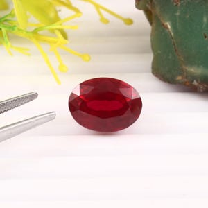 May include: A deep red, oval-cut gemstone with a faceted surface that reflects light. The stone is positioned on a white surface next to a pair of silver-toned metal tweezers, with a blurred green rock and yellow plastic plant visible nearby.