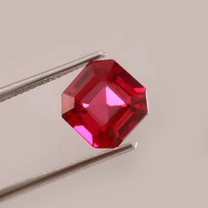 以下が含まれることがあります： 銀色のピンセットで保持された、正方形にカットされた深紅色の宝石。 宝石には、その多くのファセットと内部構造を示す、鮮やかで反射性の表面があります。 宝石は鮮やかで飽和した赤色です。