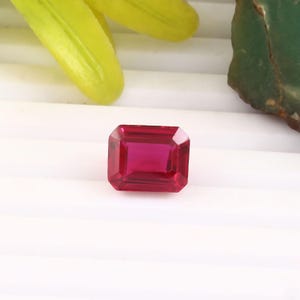 May include: A rectangular-cut ruby gemstone, displaying a rich, deep red hue. The gem has precisely cut facets and sharp edges. The ruby is set against a white backdrop, with green and yellow accents.