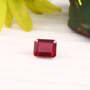 Puede incluir: Una gema rectangular, facetada, de color rojo intenso. La gema se encuentra sobre un fondo blanco, con una flor amarilla borrosa y follaje verde en el fondo. Las facetas de la gema reflejan la luz.