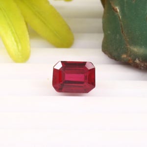 May include: A faceted, rectangular-cut, deep red gemstone. The gem has a polished surface and is set against a white background, with hints of green and yellow foliage in the background.