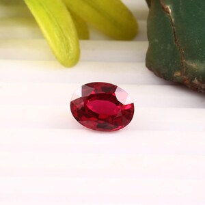 May include: An oval-cut, faceted ruby gemstone. The ruby is a deep, vibrant red color and reflects light, showcasing its facets. The gemstone is set against a white background, with hints of green and yellow foliage.