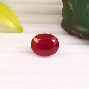 May include: A faceted, oval-shaped ruby gemstone. The gem is a deep, vibrant red color, with multiple facets catching the light. The ruby is set against a white background, with a green and yellow accent in the background.