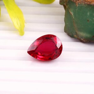 Puede incluir: Una piedra preciosa roja tallada en forma de pera con un acabado brillante y transparente. La piedra muestra tonos carmesí profundos y cortes geométricos nítidos que reflejan la luz en toda su superficie, exhibiendo un aspecto pulido.