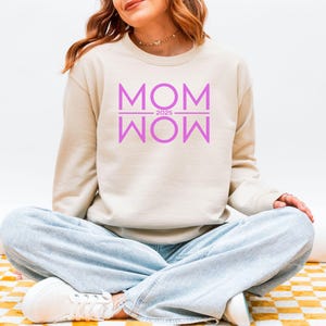 Könnte beinhalten: Beiges Sweatshirt mit dem lila Schriftzug "MOM 2025 WOW". Das langärmlige Oberteil wird mit hellblauen Jeans und weißen Turnschuhen kombiniert. Die Person sitzt im Schneidersitz.
