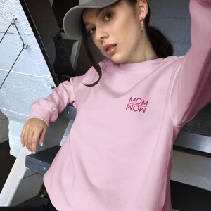 Puede incluir: Una sudadera rosa claro con la palabra "MOM" en una fuente rosa más oscura. La sudadera tiene mangas largas y cuello redondo. La modelo lleva una gorra de béisbol gris.