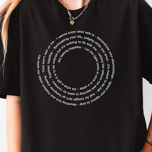 Può includere: T-shirt nera con un design circolare di testo bianco. Il testo include frasi come "Voglio sapere cosa è sicuro" e "Lascia entrare la luce di tanto in tanto". Un capo d'abbigliamento casual.
