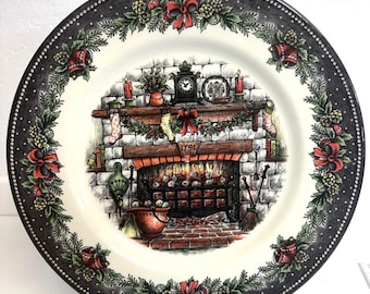 May include: A decorative plate with a Christmas scene. The plate features a detailed illustration of a fireplace with a fire burning inside. The plate has a black border with a repeating pattern of red bows, pinecones, and greenery.
