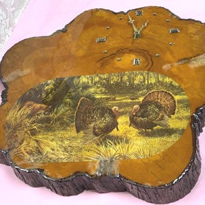 May include: A rustic wooden clock with a natural edge, featuring a wildlife scene with turkeys and a forest backdrop. The clock face has Roman numeral hour markers and decorative hands. The clock measures approximately 15 inches in diameter.