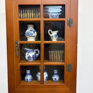 May include: A wooden display cabinet with glass doors, showcasing miniature blue and white porcelain vases, pitchers, and decorative books. The cabinet has a warm brown finish and brass hardware.
