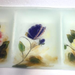 Peut inclure: Un plat de service rectangulaire en verre dépoli avec trois compartiments. Chaque section présente un motif floral peint à la main : une rose rose, un papillon violet et une rose jaune. Idéal pour servir des apéritifs ou des collations.