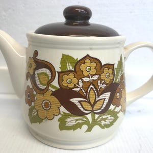 Puede incluir: Tetera de cerámica color crema con tapa y asa marrones. La tetera presenta un diseño floral en tonos marrones, amarillos y verdes. El pico y el asa son de color crema. Un artículo de cocina de estilo vintage. Mide unos 20 cm de alto.