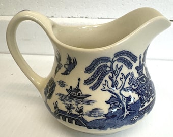 May include: Cream-colored ceramic creamer with a blue Willow pattern. The creamer features a curved handle and a spout for easy pouring. The blue pattern depicts a landscape with a bridge, trees, and buildings. A classic piece for serving cream or milk.