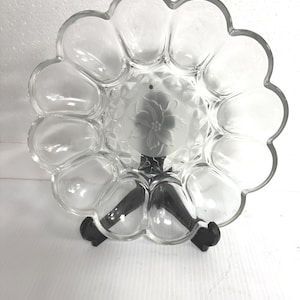 Peut inclure: Plat de service en verre transparent avec un motif floral au centre. Le plat a un bord festonné et est soutenu par un support noir. Le plat est conçu pour contenir des amuse-gueules ou des collations.