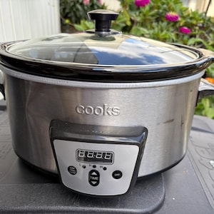 May include: A Cooks slow cooker with a stainless steel finish, black handles, and a glass lid. The digital display shows the time. The slow cooker is on a black surface.
