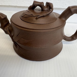 May include: Brown ceramic teapot with bamboo-style handle, spout, and lid. The lid features a bamboo branch handle and leaf details. The teapot has a round base and a cylindrical body with etched bamboo leaf designs. A traditional tea accessory.