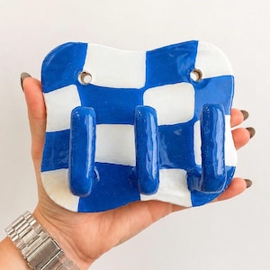K&ouml;nnte beinhalten: Eine Wandhakenleiste mit einer gl&auml;nzenden, handgeformten Keramikbasis in einem blau-wei&szlig;en Schachbrettmuster. Das rechteckige St&uuml;ck hat abgerundete Ecken, zwei kreisf&ouml;rmige Befestigungsl&ouml;cher an der Oberseite und drei hervorstehende blaue Haken.