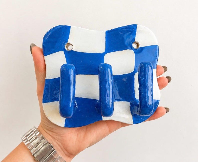K&ouml;nnte beinhalten: Eine Wandhakenleiste mit einer gl&auml;nzenden, handgeformten Keramikbasis in einem blau-wei&szlig;en Schachbrettmuster. Das rechteckige St&uuml;ck hat abgerundete Ecken, zwei kreisf&ouml;rmige Befestigungsl&ouml;cher an der Oberseite und drei hervorstehende blaue Haken.