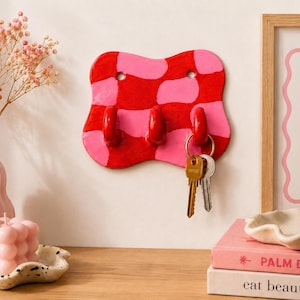 K&ouml;nnte beinhalten: Ein wandmontierter Schl&uuml;sselhalter mit wellenf&ouml;rmiger, unregelm&auml;&szlig;iger Form und einem gl&auml;nzenden rot-rosa Karomuster. Das St&uuml;ck hat drei hervorstehende Haken, von denen einer einen Metallschl&uuml;sselring mit zwei Schl&uuml;sseln h&auml;lt. Oben sind zwei L&ouml;cher.