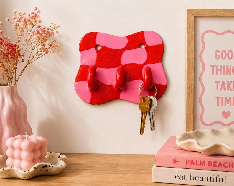 K&ouml;nnte beinhalten: Ein wandmontierter Schl&uuml;sselhalter mit wellenf&ouml;rmiger, unregelm&auml;&szlig;iger Form und einem gl&auml;nzenden rot-rosa Karomuster. Das St&uuml;ck hat drei hervorstehende Haken, von denen einer einen Metallschl&uuml;sselring mit zwei Schl&uuml;sseln h&auml;lt. Oben sind zwei L&ouml;cher.
