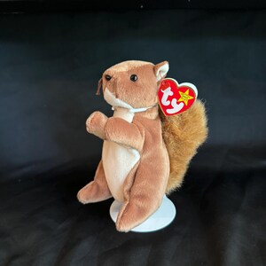 May include: A plush squirrel toy with brown fur, a white belly, and a fluffy tail. It has a red heart-shaped tag attached. The squirrel is standing on a small white base.