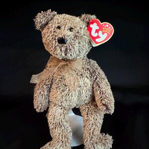 Puede incluir: Un oso de peluche marrón con un lazo de cinta alrededor del cuello. El oso tiene una nariz negra y ojos negros pequeños. Una etiqueta roja en forma de corazón con las palabras "Beanie Babies" está unida a su oreja.