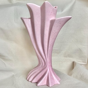 May include: A pale pink ceramic vase featuring a distinct fan-like shape with vertical, sweeping ridges that flare outward at the top and base. The surface has a subtle, speckled texture throughout, creating a matte finish on the curved structure.