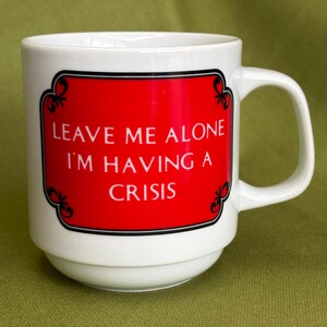 May include: A white ceramic mug featuring a rectangular red label with a black decorative border. The text on the label reads, "LEAVE ME ALONE I'M HAVING A CRISIS" in white capital letters. The mug has a rounded base and a side handle.
