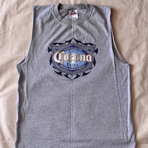 以下が含まれることがあります： 胸元にCorona Extraのロゴが青と金でプリントされたグレーのノースリーブTシャツ。ロゴには「La Cerveza Mas Fina」および「Brewed & Bottled in Mexico」という文字が装飾的な盾のデザインの中に含まれています。クルーネック仕様です。