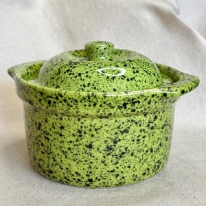 Puede incluir: Una cazuela redonda de cerámica con tapa a juego, que presenta un esmalte verde lima vibrante cubierto con un denso patrón de motas negras. El recipiente tiene dos pequeñas asas laterales integradas y un pomo central en la tapa.