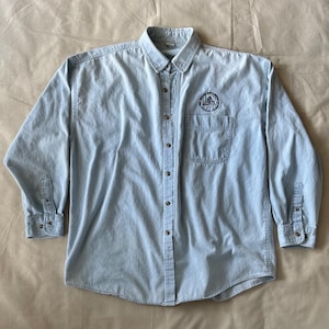 Peut inclure: Une chemise en jean bleu clair à manches longues avec une fermeture boutonnée, un col et une poche poitrine unique. La chemise présente un logo circulaire sur la poche avec le texte "ROCK AND ROLL" et "HALL OF FAME" autour d'une icône pyramidale.