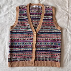 Op de afbeelding: Een mouwloos gebreid vest met een V-hals en een knoopsluiting aan de voorzijde. Het kledingstuk heeft een meerkleurig horizontaal patroon in tinten beige, marineblauw, magenta en lichtblauw, afgewerkt met een effen beige geribbelde boord.