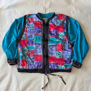 May include: A long-sleeve jacket featuring a patchwork front panel with vibrant red, teal, and purple geometric patterns. The jacket has solid teal sleeves, a black quilted trim along the front zipper, black cuffs, and a drawstring hem at the waist.