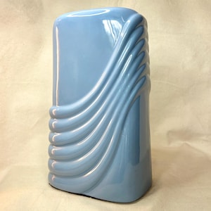 May include: A light blue ceramic vase with a smooth, glossy finish. The vase features a rectangular shape with rounded corners and a series of raised, diagonal, wave-like ridges that sweep across the front surface from the bottom left to the top right.