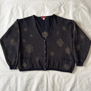 May include: A black V-neck cardigan featuring a subtle metallic gold shimmer and a repeating pattern of geometric snowflake-like motifs. The garment includes a front button closure, long sleeves with ribbed cuffs, and two front patch pockets.