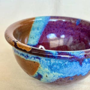 May include: A ceramic bowl featuring a glossy glaze with a blend of deep purple, vibrant turquoise, and earthy brown tones. The exterior and interior surfaces display a mottled, reactive finish with visible color transitions and a smooth, rounded rim.