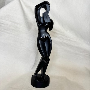 May include: A matte black sculpture of a stylized human figure standing on a circular base. The figure has an elongated form with one arm raised above the head and features subtle wavy line details on the upper torso area with some paint wear.