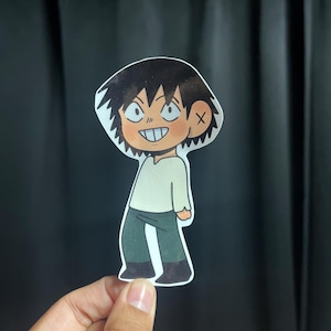 May include: A cartoon sticker of a character with spiky black hair, a cross-shaped bandage, and a wide grin. The character wears a light green long-sleeved shirt and dark green pants. The sticker is held by a hand against a dark background.