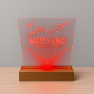 May include: A red illuminated sign with the text "GET ZAPPED MIGHTY LINKS Permanent Jewelry" on a clear acrylic panel. The panel is set in a wooden base. The sign features a lightning bolt graphic.
