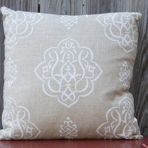 May include: Square beige throw pillow with a white geometric pattern. The pillow features a large central design and smaller corner motifs. The fabric has a textured appearance, and the pillow is placed on a red surface against a wooden fence.