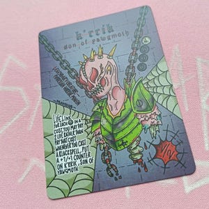 Pode incluir: Um cartão de fantasia com uma criatura grotesca com cabeça rosa, armadura verde e correntes. O cartão tem texto incluindo "K'rrik, Son of Yawgmoth" e "Lifelink". O fundo é um padrão de tijolos cinza com um design de teia de aranha.